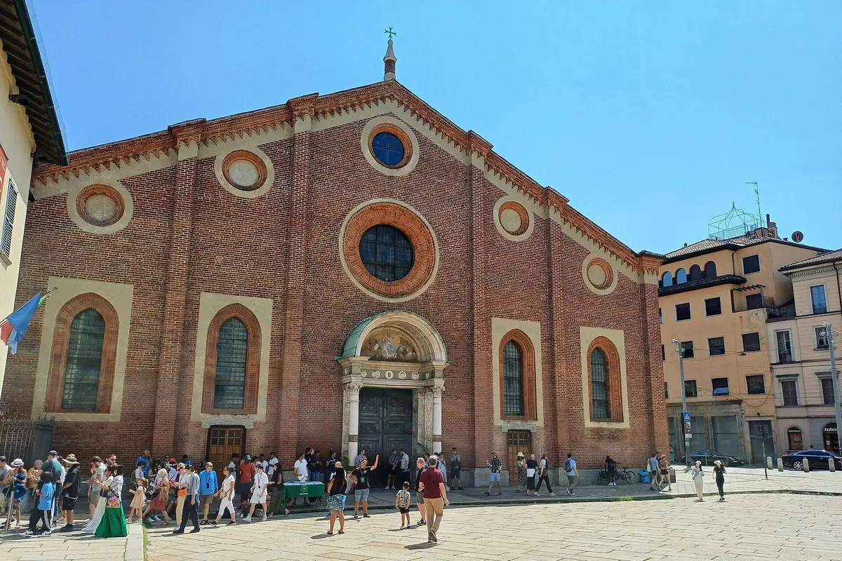 The brick facade of the church of Santa Maria delle Grazie in Milan, with a round window, a carved doorway and visitors gathered in the square